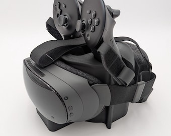 May include: A black virtual reality headset with two matching handheld controllers resting on top. The headset features a smooth front faceplate, adjustable black fabric straps, and a padded interior lining. The controllers have circular touchpads, analog sticks, and multiple buttons.
