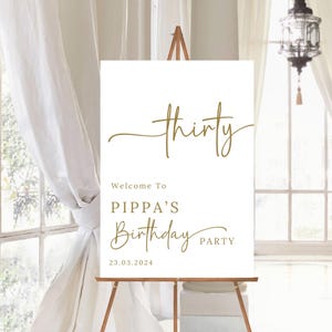 May include: A white sign on a wooden easel with gold text that reads "thirty". Below it says "Welcome To PIPPA'S Birthday PARTY" and the date "23.03.2024". The sign is in front of a window with white curtains.