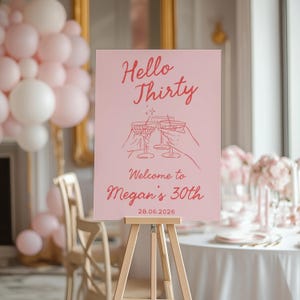 May include: A rectangular pink event sign on a wooden easel featuring the red text Hello Thirty, Welcome to Megan's 30th, and 28.06.2026. The sign displays a line art illustration of three hands holding martini glasses with a small star accent.