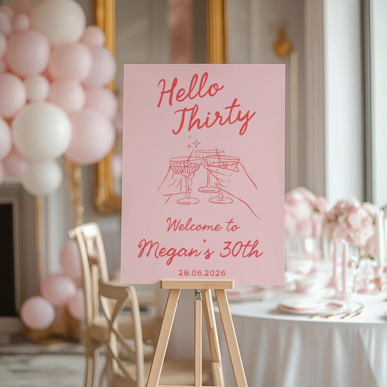 May include: A rectangular pink event sign on a wooden easel featuring the red text Hello Thirty, Welcome to Megan's 30th, and 28.06.2026. The sign displays a line art illustration of three hands holding martini glasses with a small star accent.