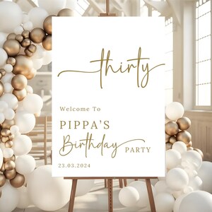 May include: A white sign on a wooden easel with gold text that reads "thirty". Below, it says "Welcome To PIPPA'S Birthday PARTY 23.03.2024". The sign is surrounded by white and gold balloons.