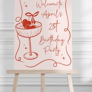 May include: A rectangular sign with a light pink background and red line art featuring a cocktail glass with cherries and a heart. The text reads: Welcome to April's 21st Birthday Party. The sign is displayed on a wooden easel with a studio logo below.