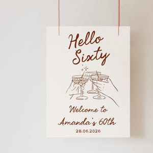 May include: A white rectangular sign with dark brown text and line art. The text reads Hello Sixty, Welcome to Amanda's 60th, and 28.06.2026. The illustration features three martini glasses being toasted, with a small star icon above them.