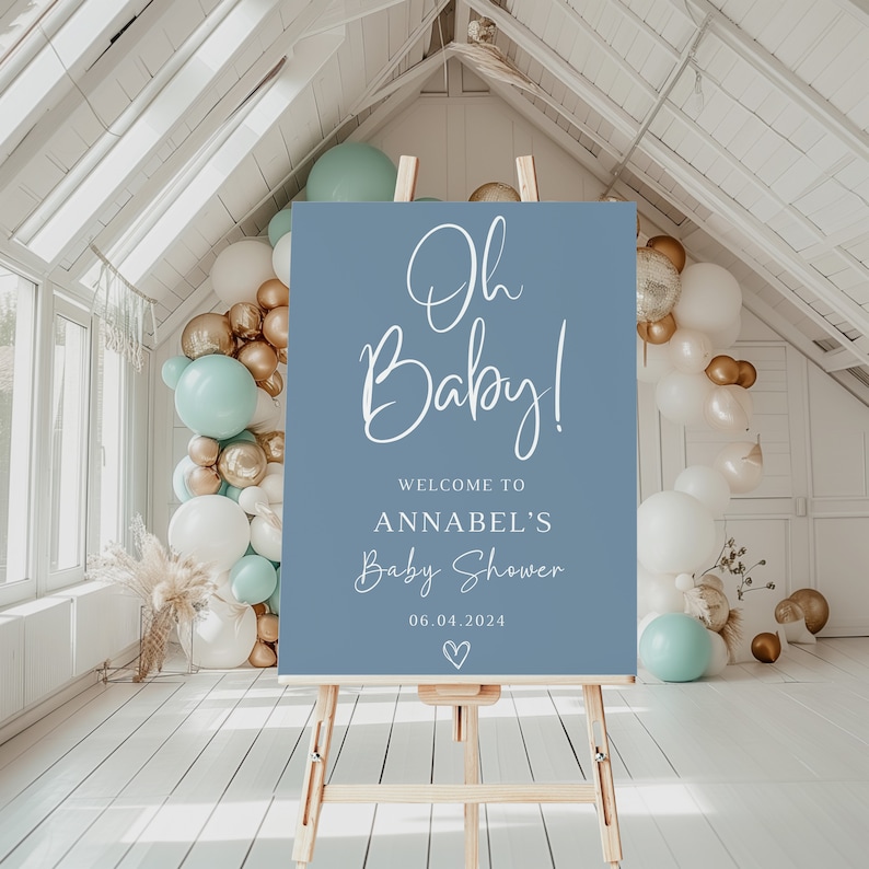 May include: A rectangular blue sign on a wooden easel featuring white text that reads Oh Baby! Welcome to Annabel's Baby Shower 06.04.2024 with a small heart icon below the date. The sign is displayed in a room decorated with white, gold, and mint balloons.