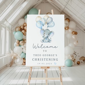 Pode incluir: Uma placa de boas-vindas retangular branca para batizado num cavalete de madeira com uma ilustra&ccedil;&atilde;o em aguarela de um elefante cinzento a segurar bal&otilde;es azuis e dourados. O texto diz Welcome to Theo George's Christening 19.05.2024 com um pequeno cora&ccedil;&atilde;o azul.