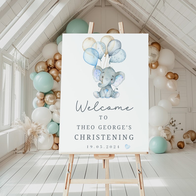 Pode incluir: Uma placa de boas-vindas retangular branca para batizado num cavalete de madeira com uma ilustra&ccedil;&atilde;o em aguarela de um elefante cinzento a segurar bal&otilde;es azuis e dourados. O texto diz Welcome to Theo George's Christening 19.05.2024 com um pequeno cora&ccedil;&atilde;o azul.