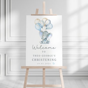 Pode incluir: Uma placa retangular branca num cavalete de madeira com uma ilustra&ccedil;&atilde;o em aguarela de um elefante cinzento a segurar bal&otilde;es azuis e beges. O texto diz Welcome to Theo George's Christening 19.05.2024 com um pequeno cora&ccedil;&atilde;o azul.