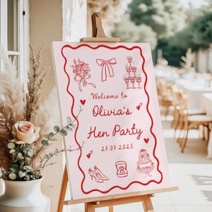 May include: A pink sign on a wooden easel, featuring a red border and illustrations. The sign says "Welcome to Olivia's Hen Party" with the date 27.07.2025. Decorative elements include a bow, champagne glasses, and a cake.