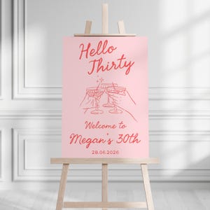 May include: A rectangular pink sign on a wooden easel featuring red text that reads Hello Thirty, Welcome to Megan's 30th, and 28.06.2026. The center graphic shows red line art of two hands holding cocktail glasses with a small star accent above.