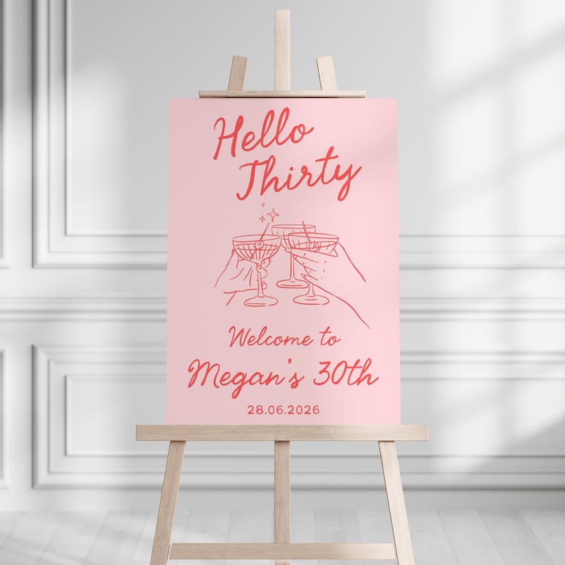 May include: A rectangular pink sign on a wooden easel featuring red text that reads Hello Thirty, Welcome to Megan's 30th, and 28.06.2026. The center graphic shows red line art of two hands holding cocktail glasses with a small star accent above.