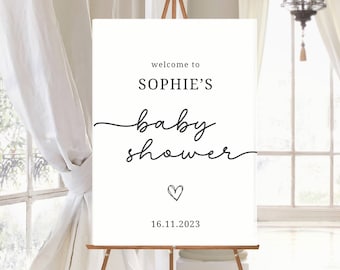May include: A white rectangular sign on a wooden easel featuring black text that reads welcome to SOPHIE'S baby shower with a small hand-drawn heart icon and the date 16.11.2023 printed in a clean, minimalist font style on the lower section.