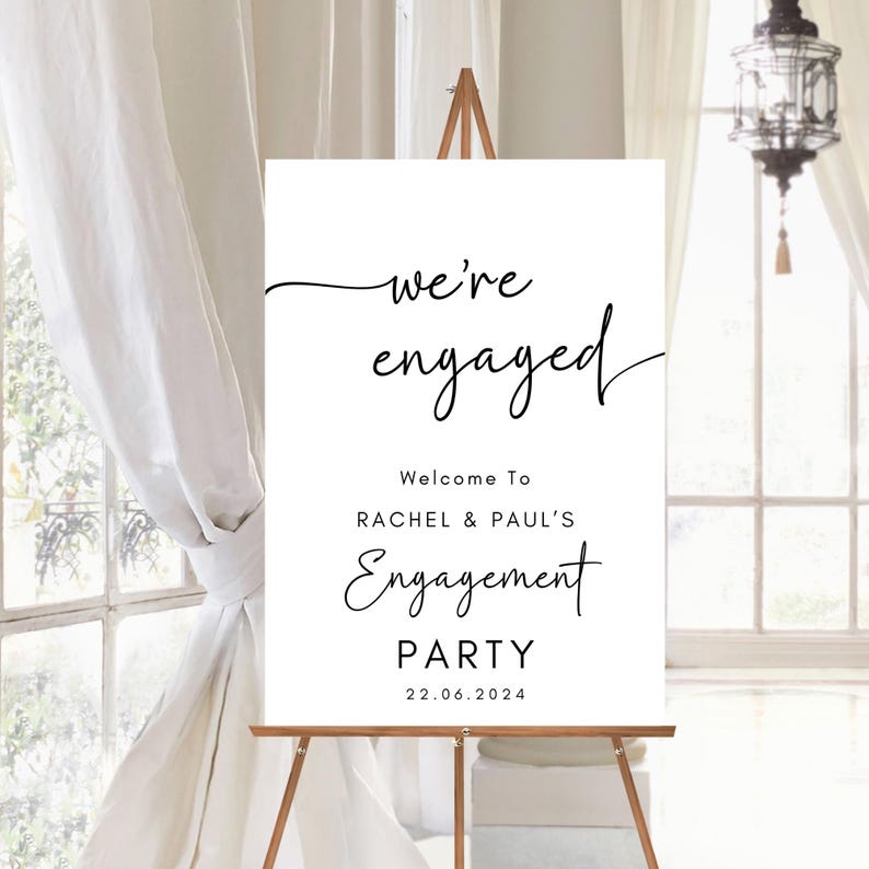 May include: A white rectangular sign on a wooden easel featuring black text that reads: we're engaged, Welcome To RACHEL & PAUL'S Engagement PARTY, and the date 22.06.2024. The design uses a mix of elegant cursive and clean sans-serif fonts.