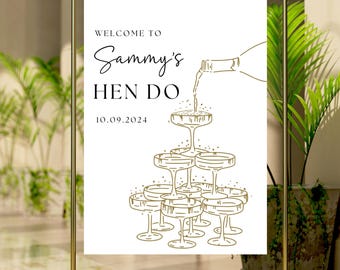 May include: White sign with gold text and illustration. The text reads "Welcome to Sammy's Hen Do 10.09.2024". The illustration shows a champagne bottle pouring champagne into a pyramid of champagne glasses.