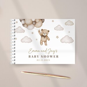 Op de afbeelding: Een spiraalgebonden gastenboek met een aquarelillustratie van een bruine teddybeer die een tros beige en bruine ballonnen vasthoudt tussen wolken en sterren. De tekst luidt Emma and Jay's BABY SHOWER 02.11.2025. Een metalen pen ligt eronder.