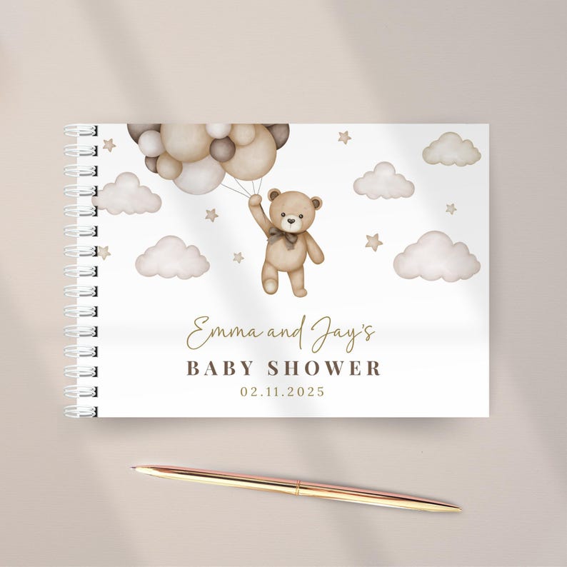 Op de afbeelding: Een spiraalgebonden gastenboek met een aquarelillustratie van een bruine teddybeer die een tros beige en bruine ballonnen vasthoudt tussen wolken en sterren. De tekst luidt Emma and Jay's BABY SHOWER 02.11.2025. Een metalen pen ligt eronder.