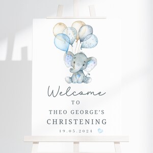 Pode incluir: Uma placa retangular branca com uma ilustra&ccedil;&atilde;o em aquarela de um elefante cinza segurando bal&otilde;es azuis e bege. O texto diz Welcome to Theo George's Christening 19.05.2024 com um pequeno &iacute;cone de cora&ccedil;&atilde;o azul abaixo da data.