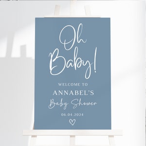 May include: A rectangular blue sign featuring white text that reads Oh Baby! WELCOME TO ANNABEL'S Baby Shower 06.04.2024 with a small hand-drawn heart icon at the bottom. The sign is displayed on a white wooden easel stand against a plain background.