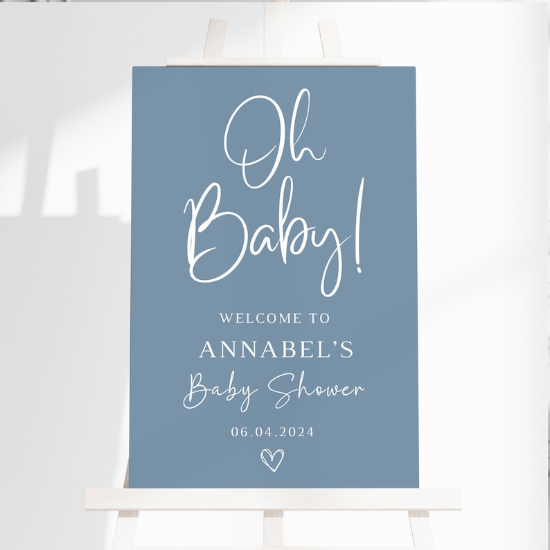 May include: A rectangular blue sign featuring white text that reads Oh Baby! WELCOME TO ANNABEL'S Baby Shower 06.04.2024 with a small hand-drawn heart icon at the bottom. The sign is displayed on a white wooden easel stand against a plain background.