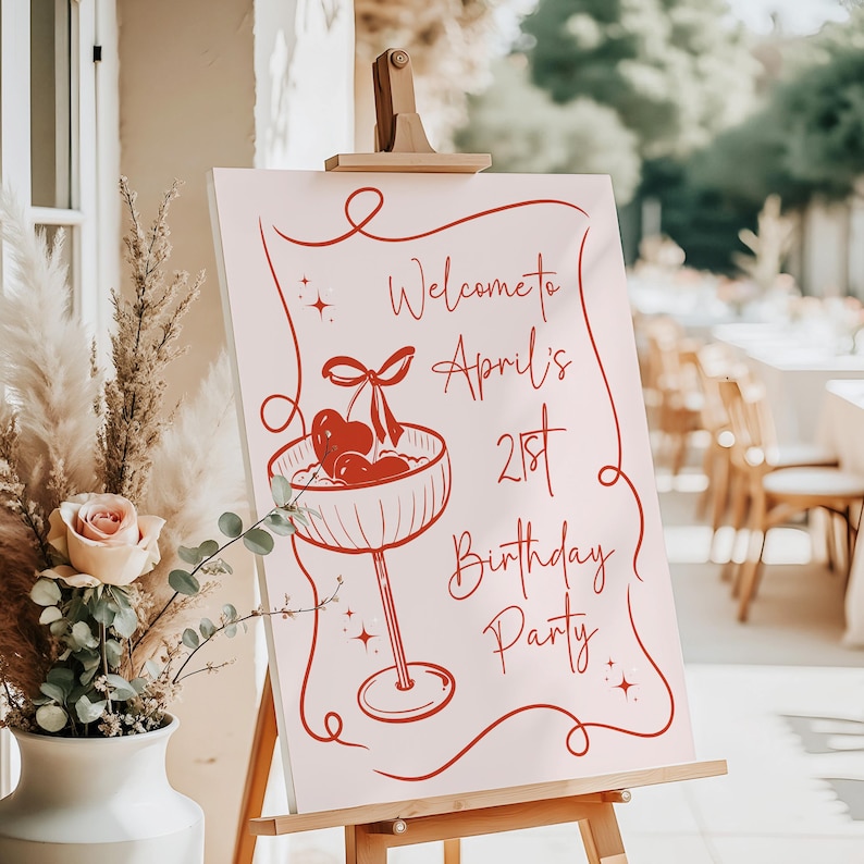 May include: A rectangular sign on a wooden easel featuring a light pink background with red line art. The design includes a cocktail glass with cherries and a bow, surrounded by a decorative border and the text Welcome to April's 21st Birthday Party.