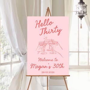 May include: A rectangular pink sign featuring red text that reads Hello Thirty, Welcome to Megan's 30th, and 28.06.2026. The center graphic displays line art of three hands holding cocktail glasses with olives, all rendered in a matching red tone.