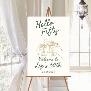 May include: A white rectangular welcome sign featuring dark green text that reads Hello Fifty, Welcome to Liz's 50th, and the date 28.06.2026. The center displays a gold line art illustration of two hands holding martini glasses with a small star.