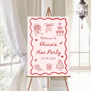 May include: A pink sign on a wooden easel, featuring red illustrations and text. The sign says "Welcome to Olivia's Hen Party" with a date. Illustrations include a bouquet, bow, champagne glasses, shoes, a ring, and a cake.