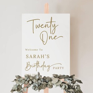 May include: A white welcome sign with gold lettering that reads "Twenty One" and "Welcome to Sarah's Birthday Party" with the date "16-03.202". The sign is on a wooden easel with greenery.
