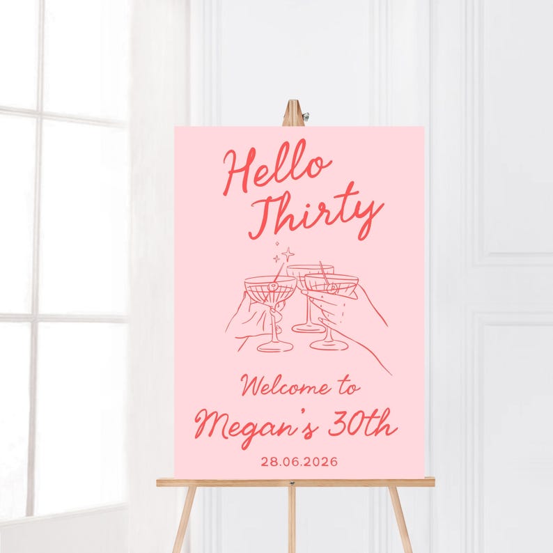 May include: A rectangular pink sign on a wooden easel featuring red text that reads Hello Thirty, Welcome to Megan's 30th, and 28.06.2026. The center displays a line art illustration of two hands holding martini glasses with a small star above them.