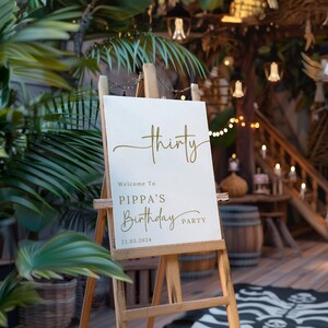 May include: A white welcome sign on a wooden easel, with gold script that reads "thirty" and "Welcome to Pippa's Birthday Party" with the date 23.03.2024. The sign is in front of a tropical background.