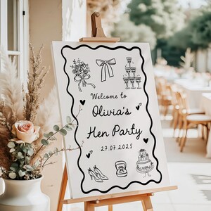 May include: A white sign on a wooden easel with black line art and text that reads "Welcome to Olivia's Hen Party 27.07.2025". The sign features illustrations of a bouquet, bow, champagne glasses, high heels, a ring, and a cake.