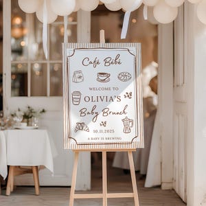 May include: A wooden easel holds a sign for a baby brunch, with the words "Café Bébé" and "Welcome to Olivia's Baby Brunch". The sign features coffee-related illustrations and the date "10.11.2025". White balloons and ribbons are visible in the background.