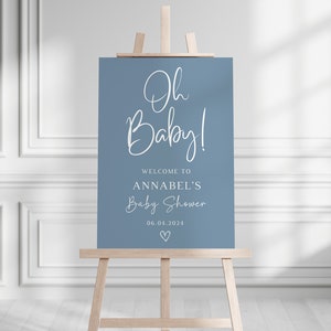 May include: A rectangular blue sign displayed on a wooden easel. The sign features white text that reads Oh Baby! WELCOME TO ANNABEL'S Baby Shower 06.04.2024 with a small heart icon at the bottom. The design uses a mix of script and sans-serif fonts.