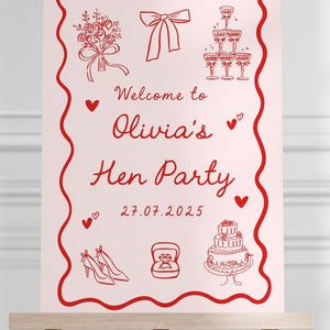 May include: A pink sign on a wooden easel with red illustrations and text. The sign reads "Welcome to Olivia's Hen Party" with drawings of a bouquet, bow, champagne flutes, shoes, a ring, and a cake. The date 27.07.2025 is shown.