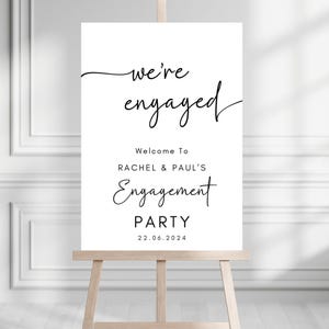 May include: A white rectangular sign on a wooden easel featuring black text that reads: we're engaged, Welcome To RACHEL & PAUL'S Engagement PARTY, and the date 22.06.2024. The design uses a combination of elegant script and clean sans-serif fonts.