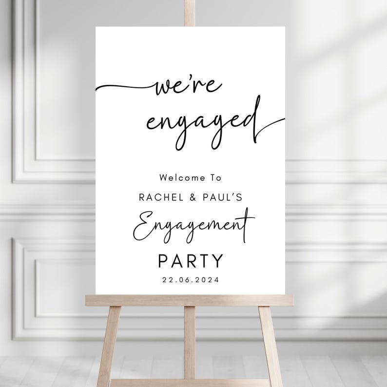 May include: A white rectangular sign on a wooden easel featuring black text that reads: we're engaged, Welcome To RACHEL & PAUL'S Engagement PARTY, and the date 22.06.2024. The design uses a combination of elegant script and clean sans-serif fonts.