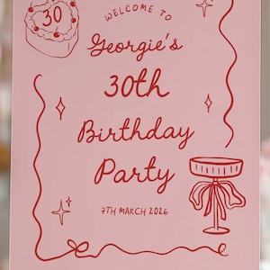 K&ouml;nnte beinhalten: Ein rosa Schild auf einer Staffelei mit roten Illustrationen und Text. Auf dem Schild steht "Willkommen zu Georgies 30. Geburtstagsparty, 7. M&auml;rz 2026." Illustrationen umfassen einen herzf&ouml;rmigen Kuchen und einen dekorativen Tisch.