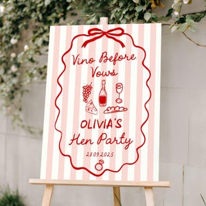 May include: A wooden easel displays a sign with pink and white vertical stripes. The sign features red text and illustrations, including the words "Vino Before Vows" and "OLIVIA'S Hen Party". The date "28.09.2025" is also visible.