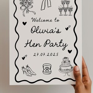 May include: A black and white sign with a decorative border, held by a hand. The sign reads "Welcome to Olivia's Hen Party" with illustrations of a bouquet, bow, champagne flutes, heels, a ring, and a cake. The date 27.07.2025 is also included.