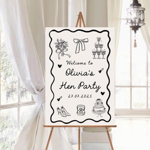 May include: A white sign on a wooden easel with black line art illustrations and text. The sign reads "Welcome to Olivia's Hen Party 27.07.2025" and includes drawings of a bouquet, bow, champagne glasses, high heels, a ring, and a cake.