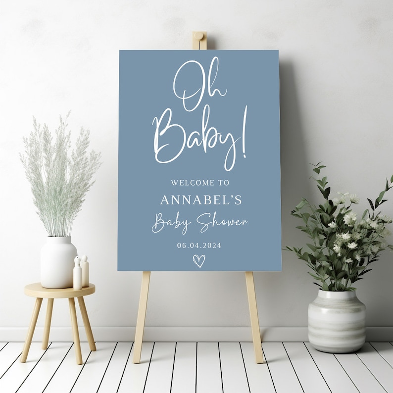 May include: A rectangular blue sign on a wooden easel featuring the white text Oh Baby! Welcome to Annabel's Baby Shower 06.04.2024 with a small hand-drawn heart icon at the bottom. The sign has a matte finish and clean, modern typography.