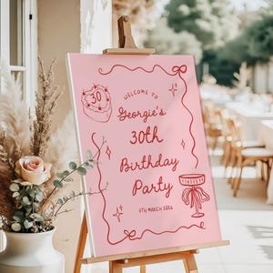 K&ouml;nnte beinhalten: Rosa Willkommensschild auf einer Staffelei f&uuml;r eine Geburtstagsfeier. Der rote Text lautet "Georgies 30. Geburtstagsparty" mit dem Datum 7. M&auml;rz 2026. Mit Illustrationen von Kuchen, Schleife und Sektglas.