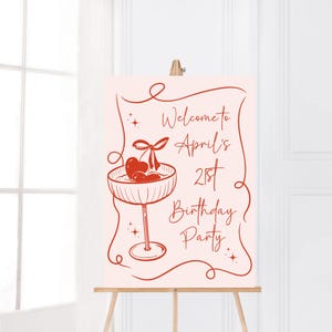 May include: A rectangular sign on a wooden easel featuring a light pink background with red line art. The design includes a cocktail glass with cherries, decorative swirls, star accents, and the text Welcome to April's 21st Birthday Party.