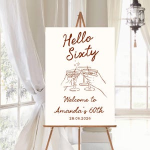 May include: A white rectangular event sign featuring brown text and line art. The text reads Hello Sixty, Welcome to Amanda's 60th, and 28.06.2026. The graphic depicts three hands holding martini glasses in a toast, accented by a small star icon.