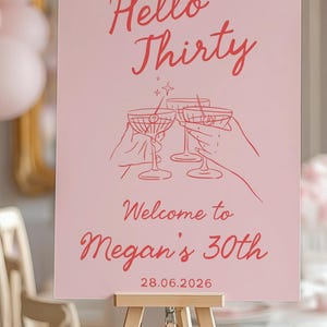 May include: A rectangular pink sign featuring red text that reads Hello Thirty, Welcome to Megan's 30th, and 28.06.2026. The center graphic displays red line art of two hands holding martini glasses in a toast, accented by small star symbols.