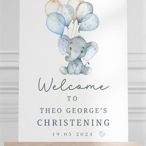 Pode incluir: Uma placa retangular branca com uma ilustra&ccedil;&atilde;o em aguarela de um elefante cinzento a segurar bal&otilde;es azuis e beges. O texto diz Welcome TO THEO GEORGE'S CHRISTENING 19.05.2024 com um pequeno &iacute;cone de cora&ccedil;&atilde;o azul junto &agrave; data.