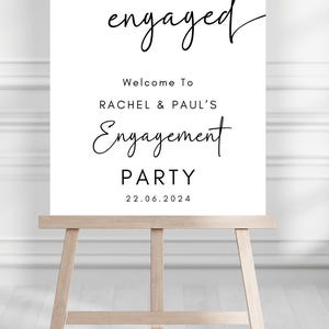 May include: A white rectangular sign on a wooden easel featuring black text that reads we're engaged, Welcome To RACHEL & PAUL'S Engagement PARTY, and the date 22.06.2024. The sign uses a combination of cursive and sans-serif typography.