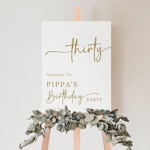 May include: A white sign on a wooden easel with gold lettering that reads "thirty", "Welcome to Pippa's Birthday Party", and the date. A garland of green leaves adorns the bottom of the sign.