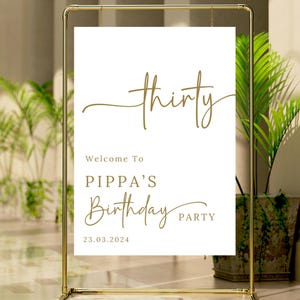 May include: A gold metal frame holds a white sign with gold text. The sign reads "thirty" in cursive, followed by "Welcome To PIPPA'S Birthday PARTY 23.03.2024". The sign is for a birthday celebration.