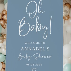 May include: A rectangular blue welcome sign for a baby shower featuring white text that reads Oh Baby! WELCOME TO ANNABEL'S Baby Shower 06.04.2024 with a small heart icon at the bottom, displayed on a light-colored wooden easel stand.