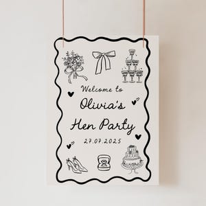 May include: A black and white sign with the text "Welcome to Olivia's Hen Party 27.07.2025". The sign features illustrations of a bouquet, bow, champagne glasses, cake, shoes, and a ring.